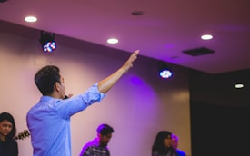 A group of people gathered indoors under soft, colorful stage lights. One person in the foreground is raising their hand, suggesting a gesture of engagement. The setting appears to be a casual or relaxed meeting or event.