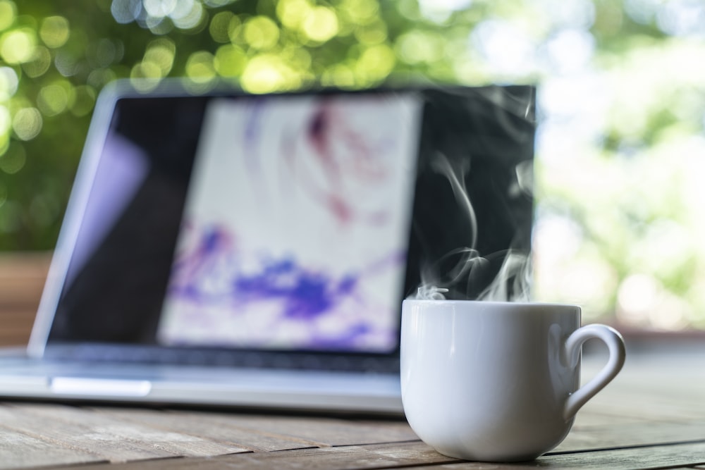 Fotografia de foco raso da caneca de cerâmica branca perto do laptop