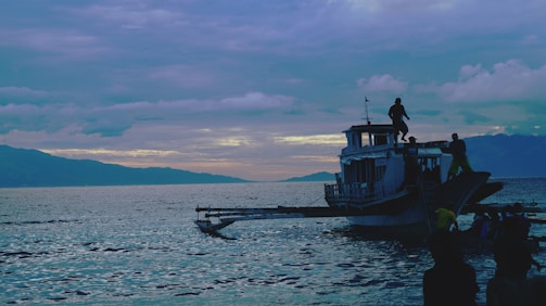 A boat filled with several people is seen on calm waters during twilight, with mountains in the background under a sky dotted with clouds. The scene conveys a sense of adventure and tranquility.