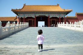 A small child wearing a pink shirt and dark pants walks toward a traditional Chinese building with ornate architecture. The structure features a prominent yellow-tiled roof and colorful patterns. Stone balustrades line the pathway leading to the entrance, where a few people are visible in the background against a clear blue sky.