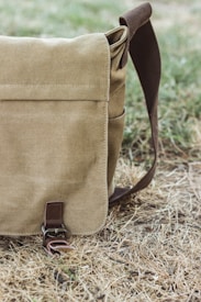 A canvas messenger bag with a flap and buckle sits on dry grass. The bag is beige with a brown strap.