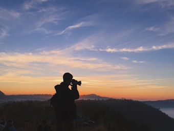 A silhouette of a person holding a camera is seen against a backdrop of a scenic sunrise or sunset. The sky is a gradient of oranges and blues, with wispy clouds scattered across. Rolling hills and a forested area are visible in the background, creating a sense of tranquility.