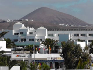 A modern white building complex is in the foreground, surrounded by palm trees. In the background, there is a large, barren mountain under a partly cloudy sky. Several flags are visible, and signs indicate businesses such as a car rental and a supermarket.