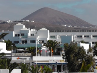 A modern white building complex is in the foreground, surrounded by palm trees. In the background, there is a large, barren mountain under a partly cloudy sky. Several flags are visible, and signs indicate businesses such as a car rental and a supermarket.