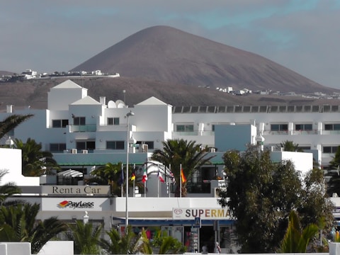 A modern white building complex is in the foreground, surrounded by palm trees. In the background, there is a large, barren mountain under a partly cloudy sky. Several flags are visible, and signs indicate businesses such as a car rental and a supermarket.