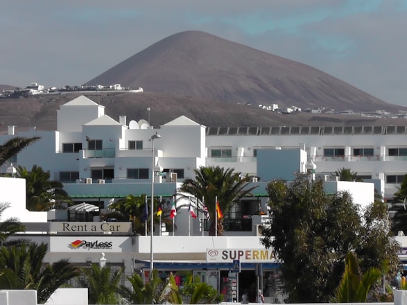 A modern white building complex is in the foreground, surrounded by palm trees. In the background, there is a large, barren mountain under a partly cloudy sky. Several flags are visible, and signs indicate businesses such as a car rental and a supermarket.
