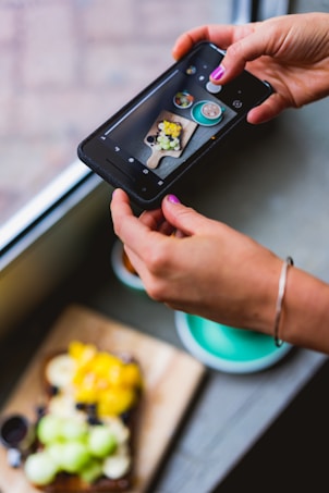 A person is holding a smartphone and taking a photo of a small meal or dessert presented on a wooden board. The phone's screen displays the focus on a dish consisting of various fruits and possibly a sauce. In the foreground, a vibrant plate and a cup are visible.