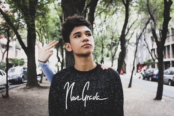 A young person is standing on a tree-lined street, surrounded by parked cars. Another person's hand is making a playful gesture near the young person's head. The scene conveys a casual urban atmosphere.