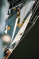 An aerial view of a sailboat with visible sails and rigging. The boat's deck is partially in sunlight with visible shadows, including a human figure. The ropes and sails create intricate patterns against the smooth, dark water below.