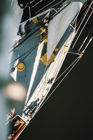 An aerial view of a sailboat with visible sails and rigging. The boat's deck is partially in sunlight with visible shadows, including a human figure. The ropes and sails create intricate patterns against the smooth, dark water below.