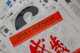 A collection of calendar pages with prominent text indicating '2018 Calendar' in bold red letters. Several pages are layered with a mix of colors, including red, black, white, and blue. There are various numbers and symbols, possibly representing dates and months, with some text in a non-Latin script.