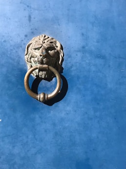 A bronze door knocker in the shape of a lion's head is mounted on a textured blue surface. The lion's facial features are detailed, and the knocker ring is held in its mouth. The background has a slightly mottled appearance with variations in the blue color.