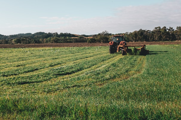 A large, green field with a red tractor is working on cutting or mowing the grass. The field is surrounded by a line of trees in the background under a partly cloudy sky. The tractor is prominently situated further back in the field, creating neat lines as it moves through the grass.