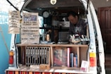A man operates a mobile coffee shop set up at the back of a van. The interior of the van is filled with coffee-making equipment including a grinder and various containers. Several signs display coffee options, blends, and prices. Books are neatly arranged on a shelf. The setting appears cozy and organized, with a focus on offering a variety of coffee experiences.