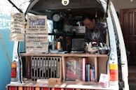 A man operates a mobile coffee shop set up at the back of a van. The interior of the van is filled with coffee-making equipment including a grinder and various containers. Several signs display coffee options, blends, and prices. Books are neatly arranged on a shelf. The setting appears cozy and organized, with a focus on offering a variety of coffee experiences.