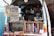 A man operates a mobile coffee shop set up at the back of a van. The interior of the van is filled with coffee-making equipment including a grinder and various containers. Several signs display coffee options, blends, and prices. Books are neatly arranged on a shelf. The setting appears cozy and organized, with a focus on offering a variety of coffee experiences.