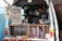 A man operates a mobile coffee shop set up at the back of a van. The interior of the van is filled with coffee-making equipment including a grinder and various containers. Several signs display coffee options, blends, and prices. Books are neatly arranged on a shelf. The setting appears cozy and organized, with a focus on offering a variety of coffee experiences.