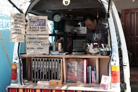 A man operates a mobile coffee shop set up at the back of a van. The interior of the van is filled with coffee-making equipment including a grinder and various containers. Several signs display coffee options, blends, and prices. Books are neatly arranged on a shelf. The setting appears cozy and organized, with a focus on offering a variety of coffee experiences.