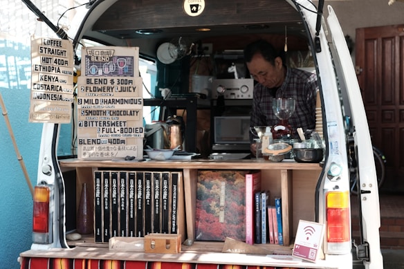 A man operates a mobile coffee shop set up at the back of a van. The interior of the van is filled with coffee-making equipment including a grinder and various containers. Several signs display coffee options, blends, and prices. Books are neatly arranged on a shelf. The setting appears cozy and organized, with a focus on offering a variety of coffee experiences.