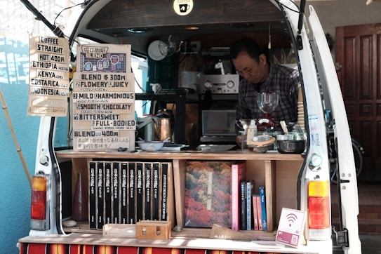 A man operates a mobile coffee shop set up at the back of a van. The interior of the van is filled with coffee-making equipment including a grinder and various containers. Several signs display coffee options, blends, and prices. Books are neatly arranged on a shelf. The setting appears cozy and organized, with a focus on offering a variety of coffee experiences.