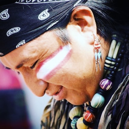 A person with long dark hair is wearing a black bandana with white patterns. They have face paint consisting of white and red stripes across the cheek. The person is adorned with colorful beaded jewelry around the neck and elaborate earrings with feather-like designs.