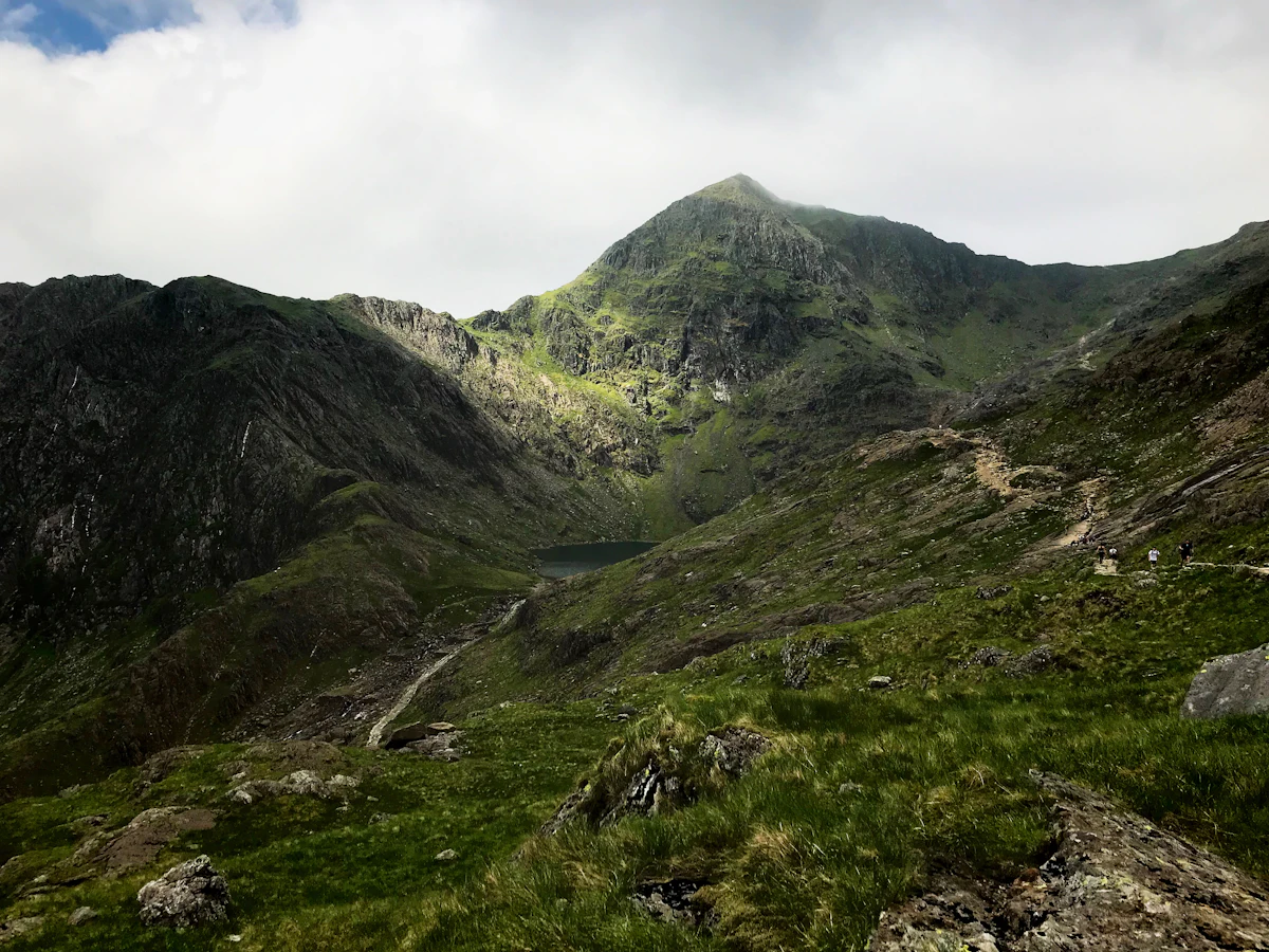 A rocky path climbing a grassy cwm toward the peak of Yr Wyddfa, with a small lake below