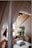 A cozy living space featuring a comfortable couch with several pillows and a white blanket draped over it. There is a potted plant with green leaves beside the couch, adding a natural element to the room. Soft, flowing white drapes hang elegantly from the ceiling, creating a serene and airy atmosphere. The room is accented by wooden textures on the walls and floor, enhancing its warm and inviting feel.