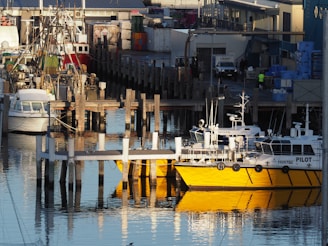 A busy harbor scene featuring several boats docked at wooden piers. The foreground includes a yellow and white pilot boat reflecting in the calm water. In the background, there are various industrial buildings and equipment, along with more boats and shipping containers. The area appears to be a working port.