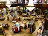 A spacious, well-lit clothing store featuring multiple tables and racks displaying a variety of garments. A large, elegant chandelier hangs from the ceiling, adding a touch of sophistication. Shoppers are seen browsing through the clothing items on display, with a mix of men's and women's apparel spread throughout the space.
