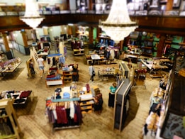 A spacious, well-lit clothing store featuring multiple tables and racks displaying a variety of garments. A large, elegant chandelier hangs from the ceiling, adding a touch of sophistication. Shoppers are seen browsing through the clothing items on display, with a mix of men's and women's apparel spread throughout the space.