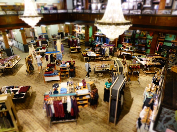 A spacious, well-lit clothing store featuring multiple tables and racks displaying a variety of garments. A large, elegant chandelier hangs from the ceiling, adding a touch of sophistication. Shoppers are seen browsing through the clothing items on display, with a mix of men's and women's apparel spread throughout the space.