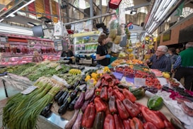 A vibrant market scene with an abundance of fresh produce displayed on a stall. Various fruits and vegetables such as peppers, bananas, grapes, and leafy greens are arranged neatly. Several people are interacting at the stall, some browsing the produce while others appear to be purchasing. The setting is lively, with an indoor market ambiance complete with lighting and other stalls in the background.