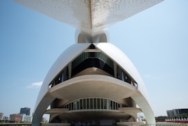 A modern architectural structure with a futuristic design, featuring smooth, curved surfaces and large, sweeping arches. The building is predominantly white with large windows, set against a clear blue sky. The lower part of the structure appears open, allowing for a view of its intricate design.