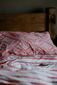 A wooden headboard with warm brown tones is partially visible behind pillows and sheets. The front pillow features a detailed red pattern with floral motifs, and the sheet displays red, coral, and white stripes. The texture of the fabric suggests a soft and comfortable bedding setup.