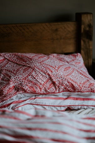 A wooden headboard with warm brown tones is partially visible behind pillows and sheets. The front pillow features a detailed red pattern with floral motifs, and the sheet displays red, coral, and white stripes. The texture of the fabric suggests a soft and comfortable bedding setup.