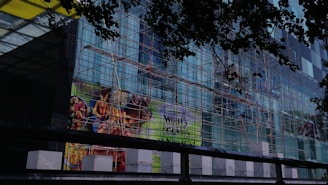 A multi-story glass building is under construction with scaffolding covering its facade. The scaffolding consists of a network of metal poles and planks. There are advertisements displayed on the glass, featuring vibrant images of food items such as fries and grilled food. The view is partially obscured by darker silhouettes of leaves, suggesting trees in the foreground.