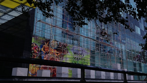 A multi-story glass building is under construction with scaffolding covering its facade. The scaffolding consists of a network of metal poles and planks. There are advertisements displayed on the glass, featuring vibrant images of food items such as fries and grilled food. The view is partially obscured by darker silhouettes of leaves, suggesting trees in the foreground.