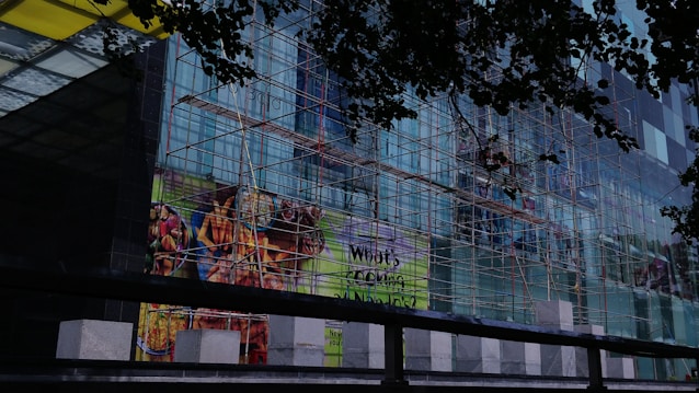 A multi-story glass building is under construction with scaffolding covering its facade. The scaffolding consists of a network of metal poles and planks. There are advertisements displayed on the glass, featuring vibrant images of food items such as fries and grilled food. The view is partially obscured by darker silhouettes of leaves, suggesting trees in the foreground.