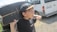 A young person is holding a skateboard over their shoulder while wearing a black cap with a casual outfit. The background includes a white van parked on a gravel path beside a grassy area.