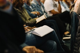 A group of people is seated closely together, with a focus on individuals holding notebooks and writing instruments. The scene suggests a learning or meeting environment.