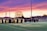 A group of graduates in caps and gowns gather on a sports field as a vibrant sunset paints the sky in shades of purple, pink, and orange. People are seen sitting on bleachers in the background.