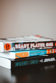 A stack of three books on a dark surface. The top book is 'Ready Player One' with a colorful cover, the middle book is 'The Hitchhiker's Guide to the Galaxy' by Douglas Adams with a white spine and blue-green text, and the bottom book is 'Armada' with a black cover and bright turquoise text.