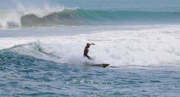 A surfer skillfully rides a wave amidst a vast expanse of ocean, demonstrating balance and agility with water splashing around. The background displays a series of rolling waves under a clear blue sky.