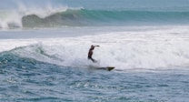A surfer skillfully rides a wave amidst a vast expanse of ocean, demonstrating balance and agility with water splashing around. The background displays a series of rolling waves under a clear blue sky.