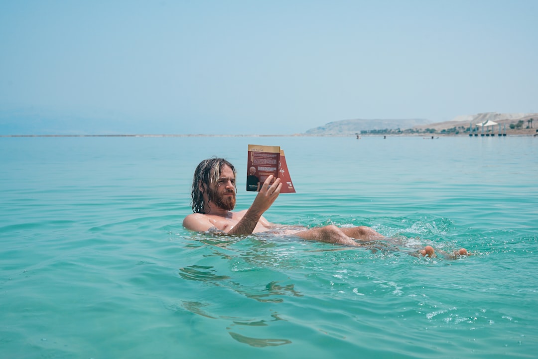 long-haired man floating in blue-green water, reading a book