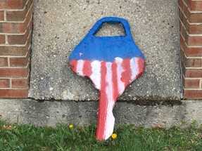 A painted object resembling a key stands upright against a rough, speckled concrete wall. The top portion of the key is painted blue, while the lower section features alternating red and white stripes. The object is placed on a bed of green grass with small yellow flowers, bordered by brick walls on either side.