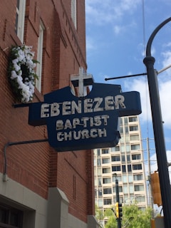 A blue sign with white lettering reads 'Ebenezer Baptist Church,' attached to the brick wall of a building. A cross is mounted on top of the sign. Below the sign is a vertical black pole, and in the background, there is a tall building with numerous windows. The sky is partly cloudy, and a white floral wreath is hung on the building.