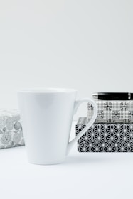 A white ceramic mug is placed next to three gift boxes with black and white geometric patterns. The background and surface are minimalistic and light in color, creating a clean and simple aesthetic.