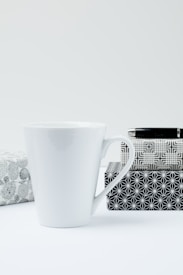A white ceramic mug is placed next to three gift boxes with black and white geometric patterns. The background and surface are minimalistic and light in color, creating a clean and simple aesthetic.