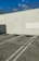 A parking area with three parking spaces is marked on an uneven, cracked asphalt surface. The wall behind the spaces is painted in a light color, featuring a small sign. The top of the wall is lined with green fencing, and a clear blue sky is visible above.